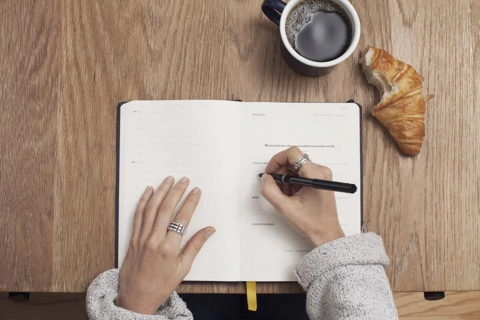 Mãos escrevendo num caderno ao lado de café e croissant