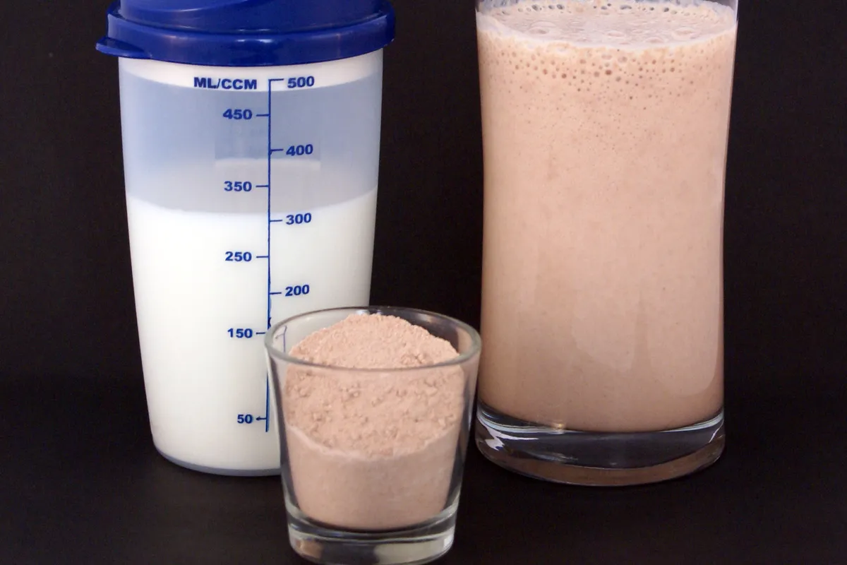 Coqueteleira com leite, copo de shake de proteína e o pó num copinho